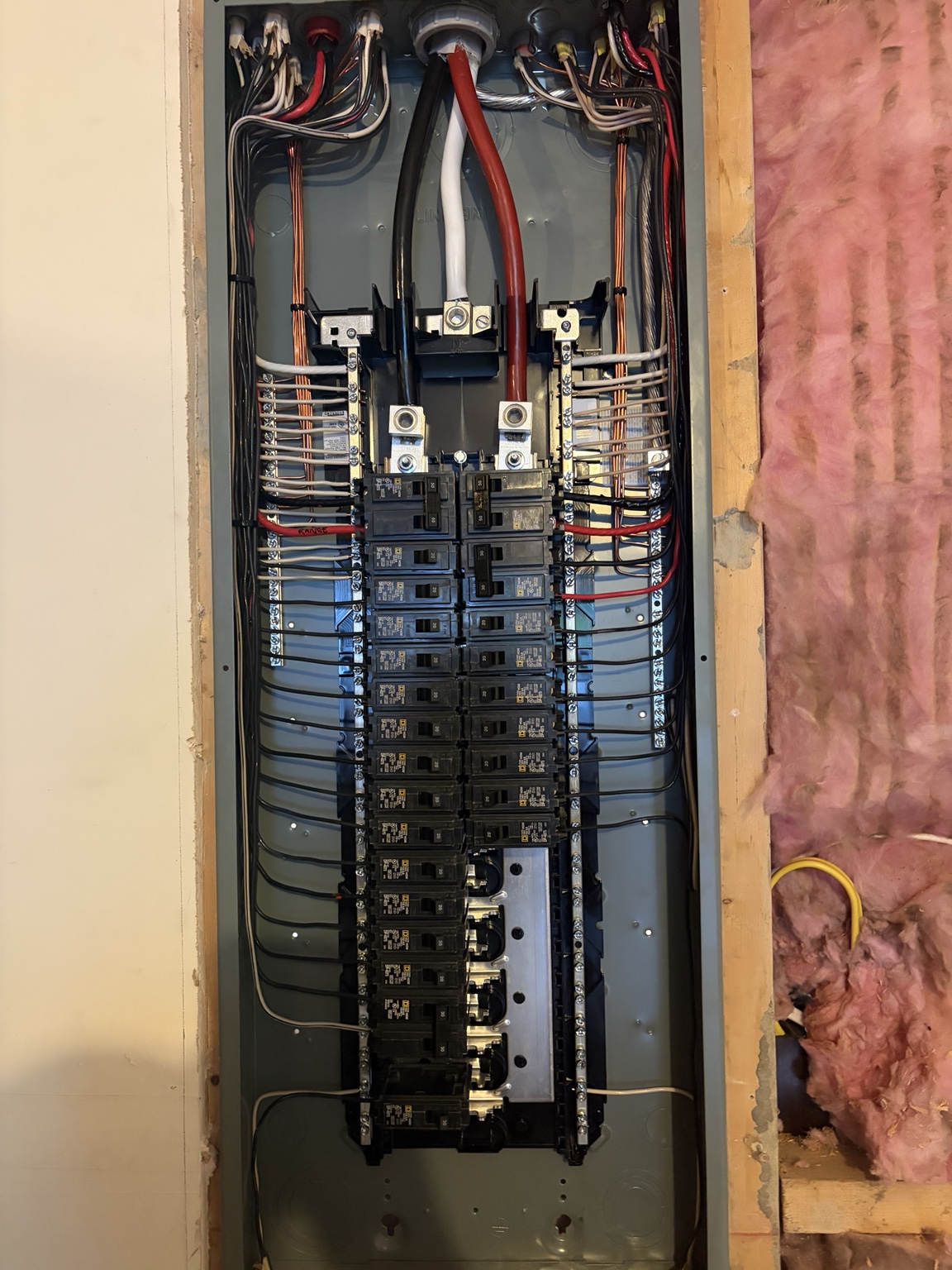 Open electrical panel with rows of circuit breakers and routed wiring between exposed framing and pink insulation