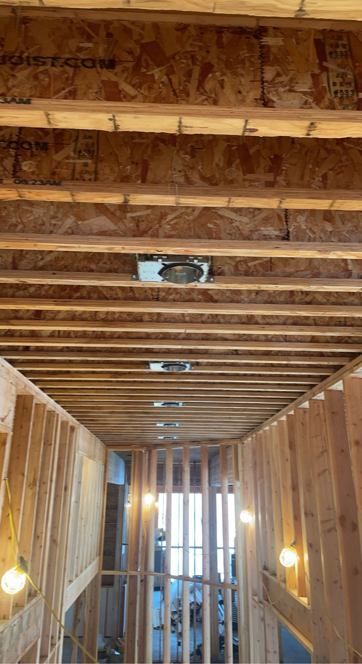 Recessed light housings mounted between exposed ceiling joists above a framed room with temporary work lights