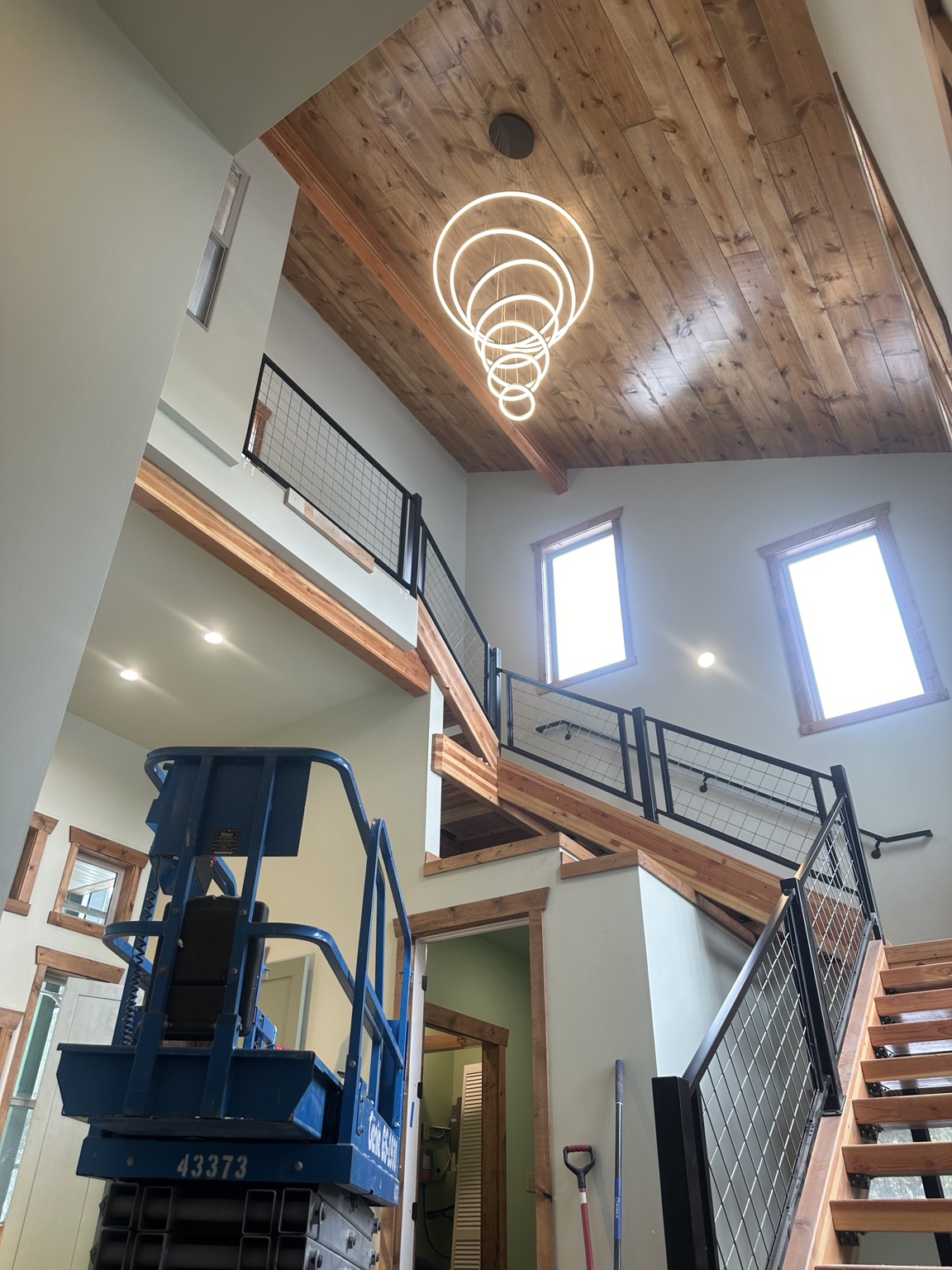 Upward view of an illuminated multi-ring chandelier beneath a wood ceiling, with open stairs, metal railings, and a blue lift below